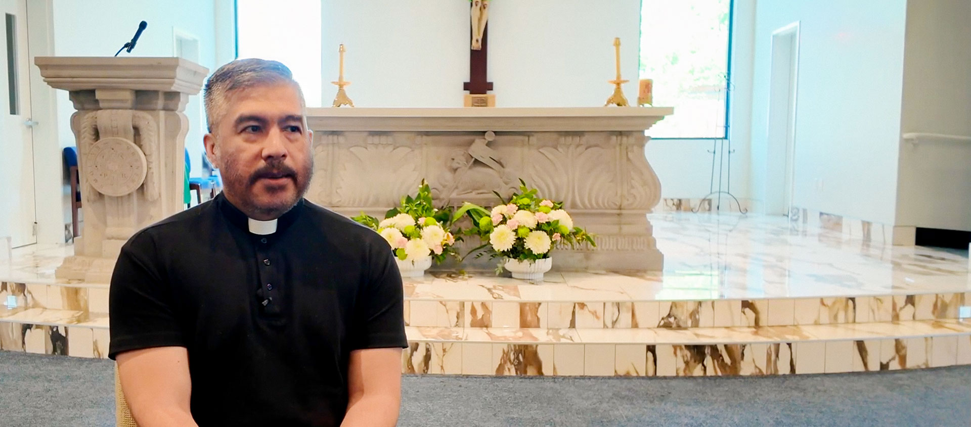 Father Edgar sits inside a church, in front of a stone altar adorned with flowers and candles
