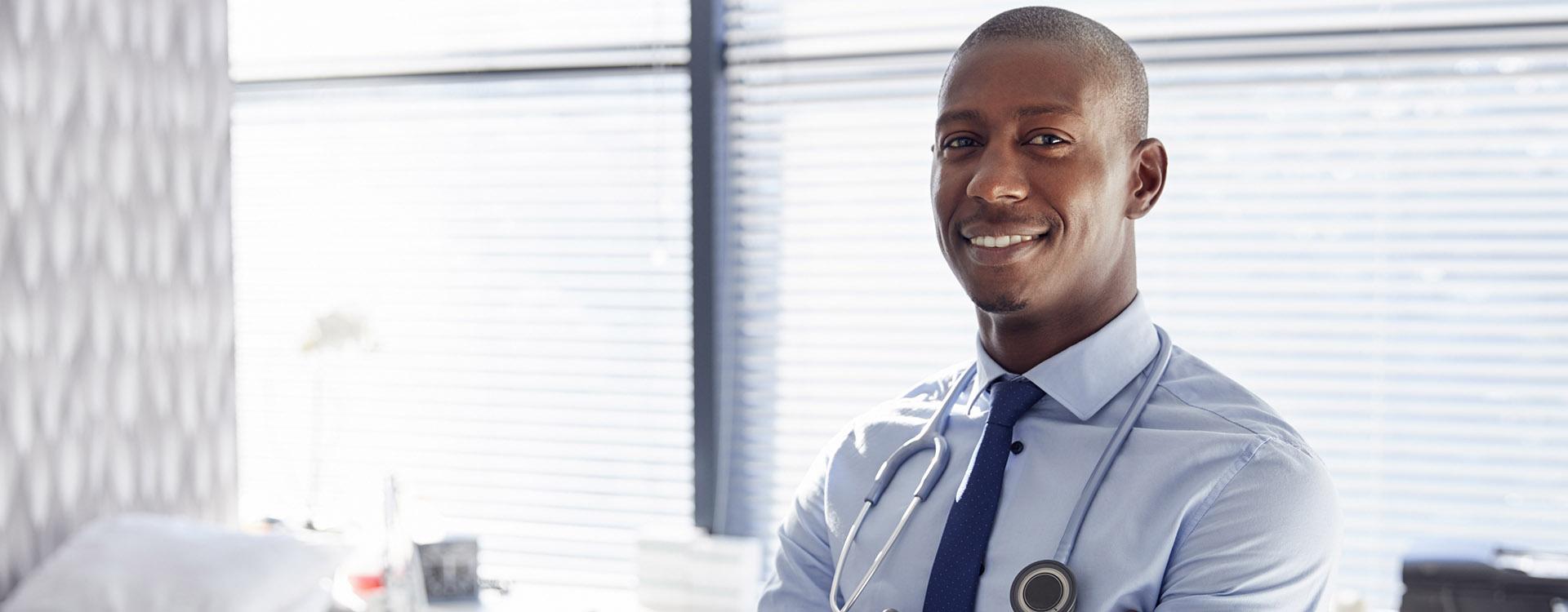 Portrait Of Smiling Male Doctor With Stethoscope Standing By Desk In Office