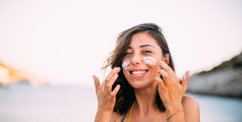 A woman outside at a beach applying sunscreen to her cheeks.