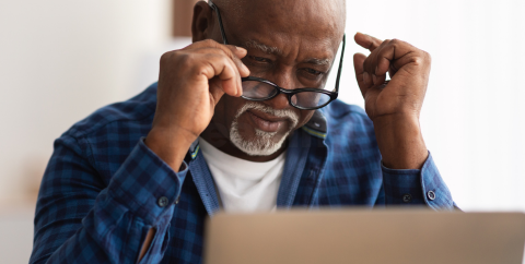 an older Black man adjusts his glasses as he looks at a laptop screen.