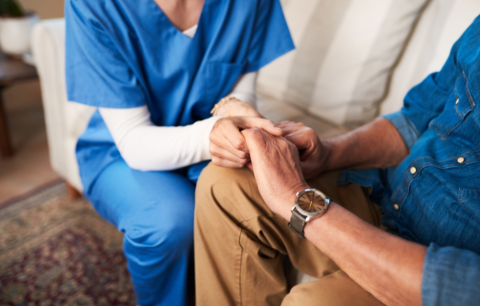 A nurse in scrubs holding the hands of an older man. The pair are sitting on a couch angled to face each other.