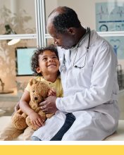 A young boy smiles and holds a teddy bear and sits next to a doctor who has his arm around the boy in a friendly manner.